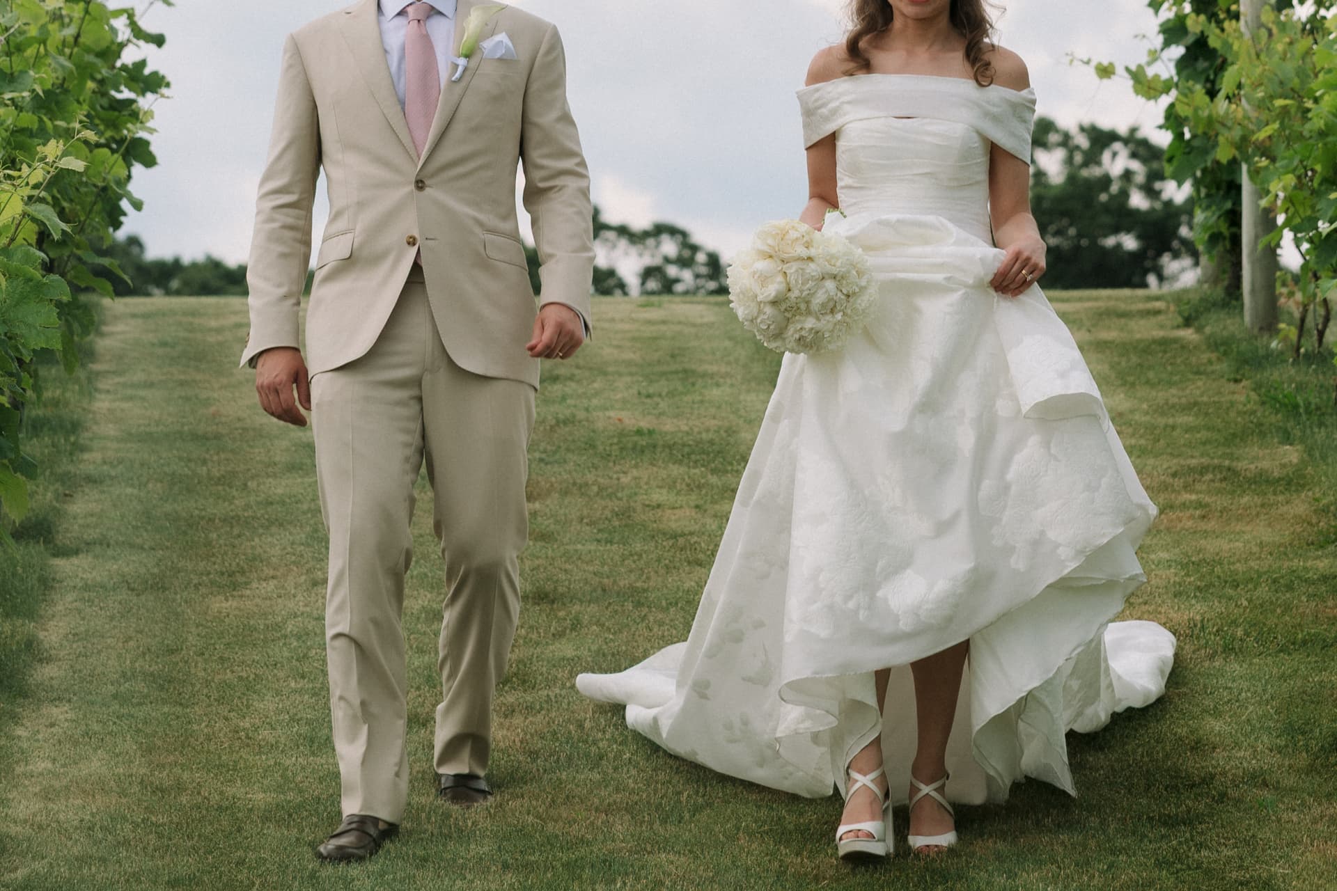 Bride and groom walking together through a grassy vineyard path